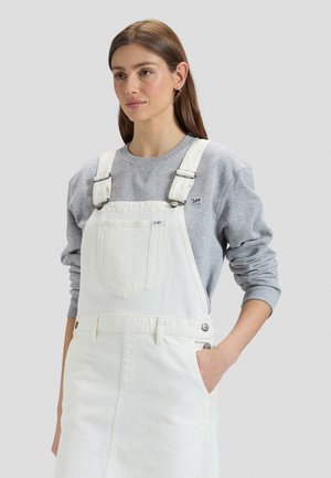 Tânără cu păr lung și castaniu, purtând un hanorac gri Lee și o rochie din denim albă, cu mâna în buzunar, pe fundal neutru.