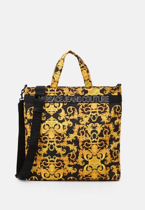 Czarna torba typu tote z ozdobnym złotym wzorem barokowym, z logo "Versace Jeans Couture" na górze, wyposażona w krótkie uchwyty i regulowany pasek.
