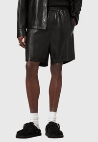 Schwarze Lederhosen mit glatter Textur, elastischem Bund und Seitentaschen. Kombiniert mit schwarzen Slipp-ons und weißen Socken.