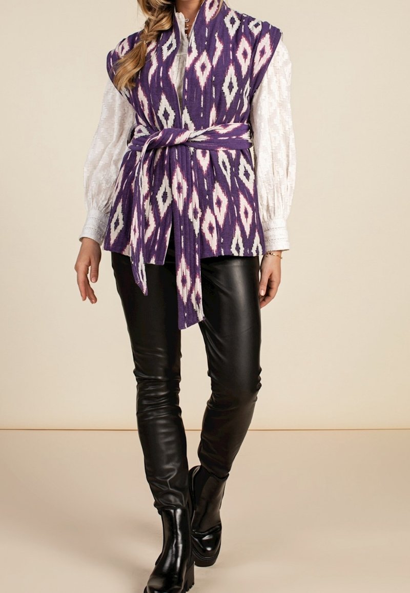Femme portant un gilet sans manches violet et blanc à motifs avec ceinture, par-dessus un chemisier blanc à manches longues, un pantalon en cuir noir et des bottes noires.