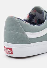 Mint green canvas sneaker with white side stripe, padded collar, and notable Vans logo tag in red on the heel. Floral lining visible inside.
