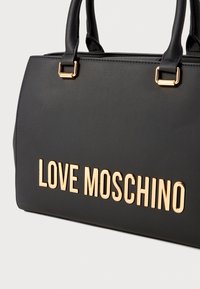 Mustmust mustkott, mis on valmistatud siledast sünteetilisest materjalist, millel on kullatoonilised metalldetailid ja eespool on väljaspool selgelt nähtav "LOVE MOSCHINO" logo.