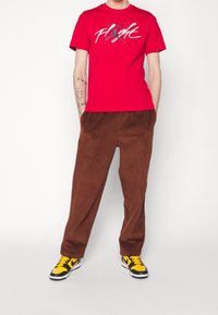 Punainen puuvillainen t-paita, jossa on "FLIGHT" -grafiikka, yhdistettynä ruskeisiin corduroy-housuihin ja musta/keltaisiin lenkkareihin, seisoo valkoista taustaa vasten.