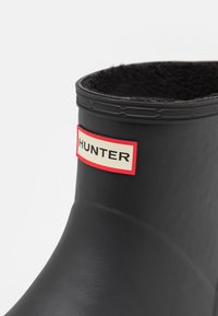 Schwarze Gummiregenstiefel mit strukturierter Oberfläche, runder Oberseite und einem roten und beigen "HUNTER"-Logoschild an der Seite.
