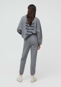  kvinde med langt brunt hår iført en grå træningsdragt med sorte håndskrevne navne på ryggen, iført hvide støvler, stående på en ensfarvet lys baggrund.