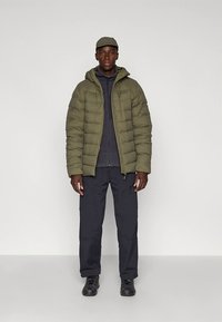 Maslinasto zelena jakna na preklop s patentnim zatvaračem i kapuljačom; ispod je slojevita siva dukserica. Plave hlače i crna obuća upotpunjuju odjevnu kombinaciju.