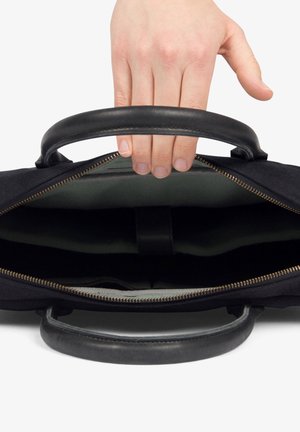 Black fabric bag with leather handles, zip closure, and green interior lining. Hand reaching into bag, showing spacious design.
