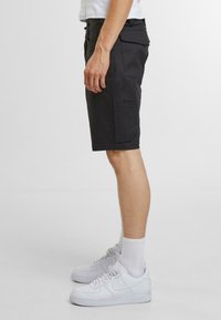 Pantaloni cargo neri realizzati in tessuto resistente, con tasche laterali e taglio dritto. Indossati con sneakers bianche e calzini alla caviglia.
