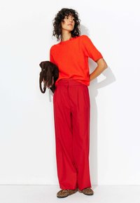 Pantalons plissés rouges taille haute associés à un pull en tricot à manches courtes orange vif. Les accessoires comprennent un sac à franges marron et des chaussures à imprimé léopard.