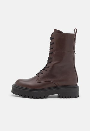 Botte de combat en cuir marron foncé avec semelle épaisse noire à crampons et lacets noirs, équipée de coutures latérales et d'une languette de traction arrière.