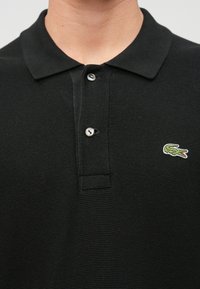 Polo noir en tissu texturé, avec un col, deux boutons argentés et un petit logo de crocodile brodé en vert.