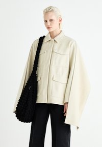 Beige oversized ullen kappejakke med fire frontlommer, kombinert med en teksturert svart skulderveske. Stylet med svarte bukser.