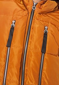 Vaatteeksi pehmustettu oranssi takki, jossa on huppu, mustat vetoketjun yksityiskohdat ja kaksi ulkoista vetoketjullista taskua rinnassa. Silkkinen polyesterimateriaali.