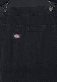 Černé lacláče vyrobené z odolné tkaniny mají přední kapsu s viditelným logem Dickies, kovové součásti a nastavitelné šle.
