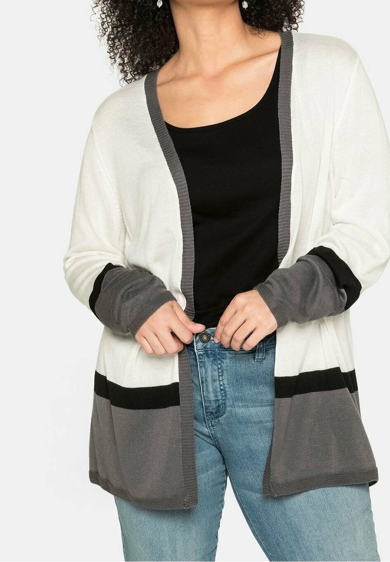 Femme portant un cardigan bicolore blanc, gris et noir sur un haut noir et un jean bleu, ajustant les bords du cardigan avec ses mains.