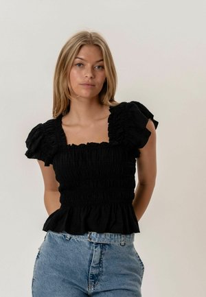 Młoda kobieta o blond włosach ubrana w czarną marszczoną bluzkę z bufkami i wysokim stanem jasnoniebieskich dżinsów na jednolitym tle.