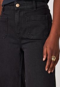 Primo piano di una persona che indossa pantaloni in denim neri con tasche frontali, un bottone dorato e una mano con un anello dorato testurizzato appoggiata accanto alla coscia.