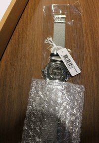 Edelstahl-Armbanduhr mit Mesh-Armband, in Kunststoff eingekapselt. Verfügt über ein klares Zifferblatt und einen schwarzen Akzent. Enthält ein Preisschild.