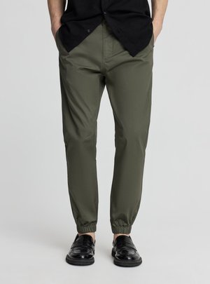 Uomo che indossa pantaloni jogger affusolati verde oliva con polsini elastici abbinati a mocassini neri e una camicia nera, in piedi con le mani in tasca.