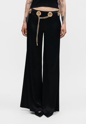 Čierne nohavice s širokými nohavicami s veľkými zlatými okami a zlatým reťazovým opaskom, nosené s čiernymi špicatými topánkami a viditeľnými tetovaniami na rukách.