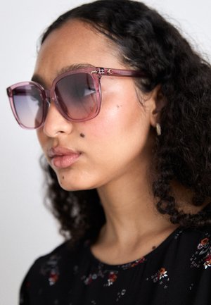 Ochelari de soare mari, supradimensionați, cu un cadru roz translucid și lentile cu degrade. Prezintă accesorii de culoare aurie și o formă geometrică.