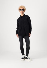 Oversized košulja s uzorkom crnog leoparda, uparena s crnim uskim trapericama i crnim tenisicama s bijelim potplatima. Model nosi velike sunčane naočale.