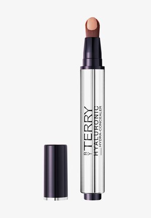 Hyaluronische concealer in een zilveren tube met een donkerpaars dop, met een schuine borstelpunt en een nude-kleurig product aangebracht.