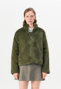 Groene nepbont jas, met rits aan de voorkant, hoge kraag, relaxte pasvorm, gestructureerde stof, lange mouwen, gedragen over een grijs geruit rokje.