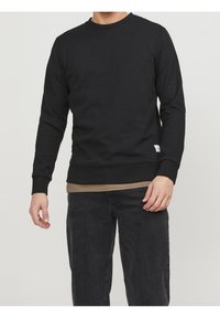 Felpa nera a collo rotondo realizzata in tessuto testurizzato, con maniche lunghe e una piccola toppa con logo sull'orlo inferiore. Indossata sopra una camicia color beige.