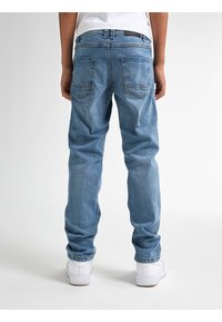 Pantalones vaqueros de mezclilla de color azul claro con un corte relajado, costuras visibles, dos bolsillos traseros y una etiqueta negra en la cinturilla, combinados con zapatillas blancas.