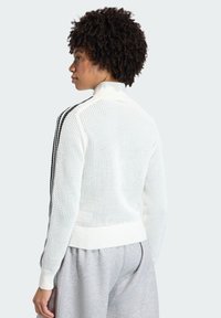 Bijeli pleteni džemper s visokim ovratnikom, s teksturiranim mrežastim uzorkom i crnim bočnim prugama. Nošen s sivim hlačama.