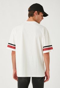 Camiseta blanca de manga corta con rayas rojas y negras en los puños. Diseño casual, textura suave, corte relajado y costuras visibles.