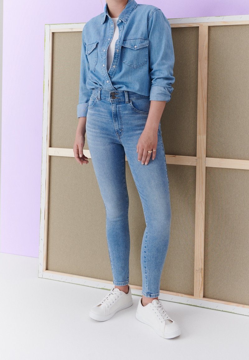 Camisa de mezclilla azul clara con dos bolsillos en el pecho, anudada en la cintura, combinada con jeans azul claro de cintura alta y zapatillas blancas.