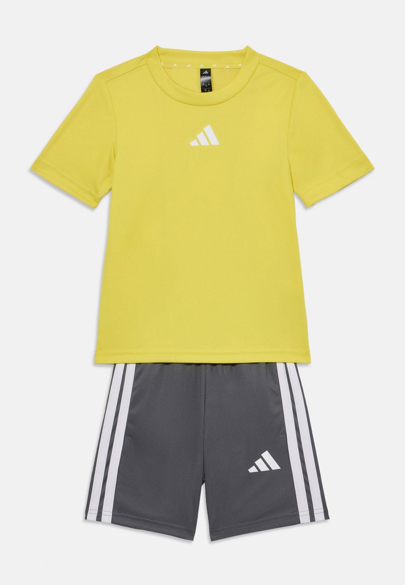 Camiseta deportiva amarilla con un logo blanco, combinada con pantalones cortos grises que presentan rayas blancas y un logo. Textura suave, diseño moderno.