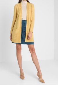 Cardigan giallo a maglia con apertura frontale, due tasche anteriori, indossato sopra un dolcevita beige e una gonna in denim blu con dettagli a bottoni; tacchi beige.