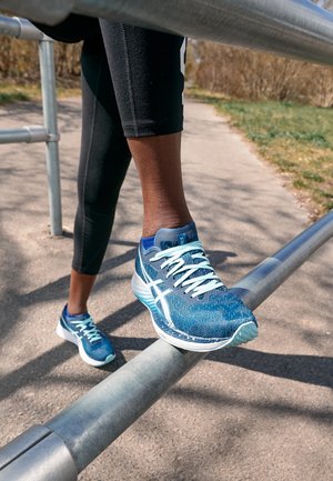 Persona con zapatillas azules de correr estirando la pierna sobre una barandilla de metal al aire libre en un camino soleado con vegetación cercana.