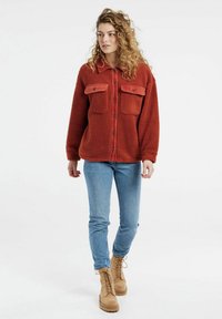 Rust-colored fleece jacket with button-up front, two chest pockets, and a soft texture. Worn with blue jeans and tan lace-up boots.