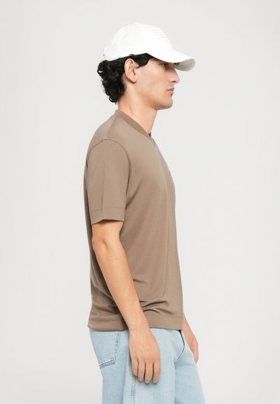 Homme en chemise beige à manches courtes, jeans bleu clair et casquette blanche, se tenant de profil contre un fond blanc uni.