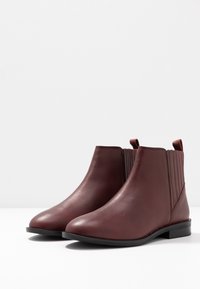 Par de suaves botas de tobillo de cuero rojo oscuro con paneles laterales elásticos y tacones negros bajos sobre una superficie blanca.