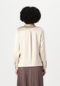 Beige satiinitoppi, jossa on kauluksellinen pääntie, pitkät hihat ja laskostus yksityiskohtana takana. Sujuva textura ja minimalistinen muotoilu.