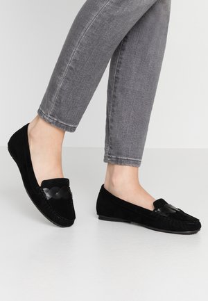 Mocassins en daim noir avec une bande texturée, bout arrondi et semelle plate. Portés avec un jean gris, illustrant un style décontracté.