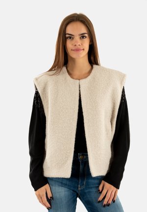 Gilet sans manches beige clair en tissu texturé, avec un col rond et une ouverture à l'avant, porté par-dessus un haut à manches longues noir.