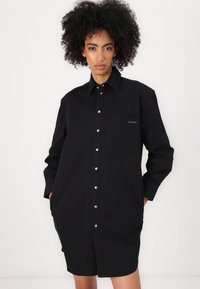 Abito camicia nero in cotone con chiusura frontale a bottoni, maniche lunghe, tasca sul petto e bottoni color argento. Design semplice e taglio sartoriale.