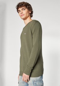 Chandail en tricot vert olive avec un col en V, des poignets et un ourlet côtelés. Présente un petit logo sur la poitrine. Le tissu a un motif texturé.