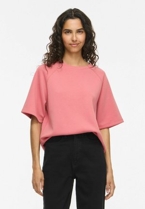 Donna con capelli lunghi e ricci che indossa una camicia rosa a maniche corte larga e pantaloni neri a vita alta, in piedi davanti a uno sfondo bianco semplice.