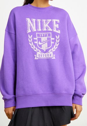 Person iført en lilla Nike sweatshirt med hvid tekst og logo "NIKE 1972 Oregon", kombineret med et sort skørt, beskåret ved skuldrene.