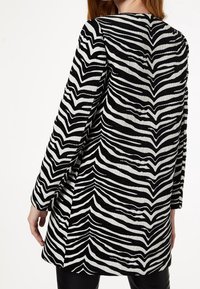 Mujer vistiendo una chaqueta de manga larga con estampado de cebra en blanco y negro, vista desde atrás contra un fondo claro y liso.