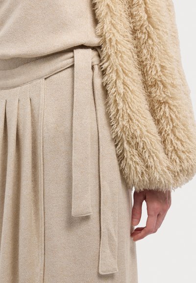 Beiges Strickoberteil mit einem flauschigen, strukturierten Ärmel. Der beige Rock mit Gürtel hat ordentliche Falten und einen subtilen Schimmer.
