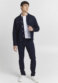 Dark denim jacket with front pockets and button closure, paired with dark denim jeans and white sneakers. Model stands against a light background.