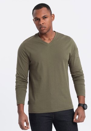 Camisa de manga larga verde oliva con cuello en V, hecha de una mezcla de algodón, que presenta un ajuste relajado y una etiqueta en el dobladillo izquierdo.